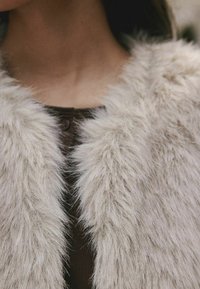 Chaleco de piel sintética en beige claro con un acabado suave y texturizado. Presenta un cuello redondo y botones en la parte delantera.