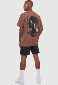 Brun grafisk t-shirt med en sort silhuet og kæde design på ryggen. Sammen med mørke shorts og hvide sneakers.