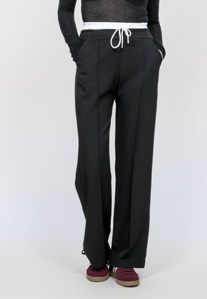 Personne portant un pantalon large noir avec un cordon blanc, un haut à manches longues noir, les mains dans les poches, et des baskets bordeaux avec des semelles beiges.