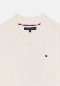 Pull en tricot torsadé crème avec un col en V, arborant une petite patte de logo Tommy Hilfiger. Col et poignets côtelés avec des accents rouges et bleus discrets.