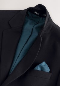 Veste de costume noire avec une doublure à motif turquoise et une pochette turquoise assortie pliée dans la poche poitrine.