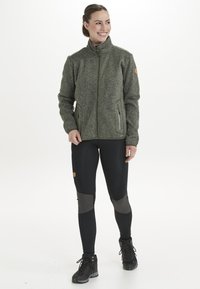 Hiilikuitutuulettimella varustettu fleece-takkia, jossa on korkea kaulus ja vetoketjullinen etuosa, yhdistetty mustiin leggingseihin, joissa on harmaat polvipatchesit, ja mustiin saappaisiin.