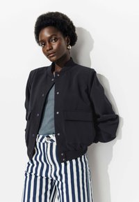 Veste bomber noire avec col côtelé, deux poches latérales, portée par-dessus un haut teal et assortie à un pantalon rayé bleu marine et blanc.