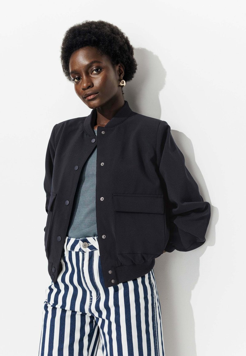 Veste bomber noire avec col côtelé, deux poches latérales, portée par-dessus un haut teal et assortie à un pantalon rayé bleu marine et blanc.