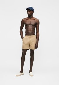 Άνδρας χωρίς πουκάμισο, φοράει μπεζ σορτς, λευκά παπούτσια slip-on και μπλε σκούρο καπέλο του μπέιζμπολ, στέκεται με τα χέρια στις τσέπες μπροστά σε απλό φόντο.