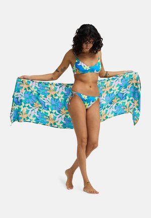 Vrouw in een blauwe bikini met bloemenpatroon die een bijpassende grote bloemenhanddoek wijd uitspreidt, blootsvoets staand op een effen lichte achtergrond.