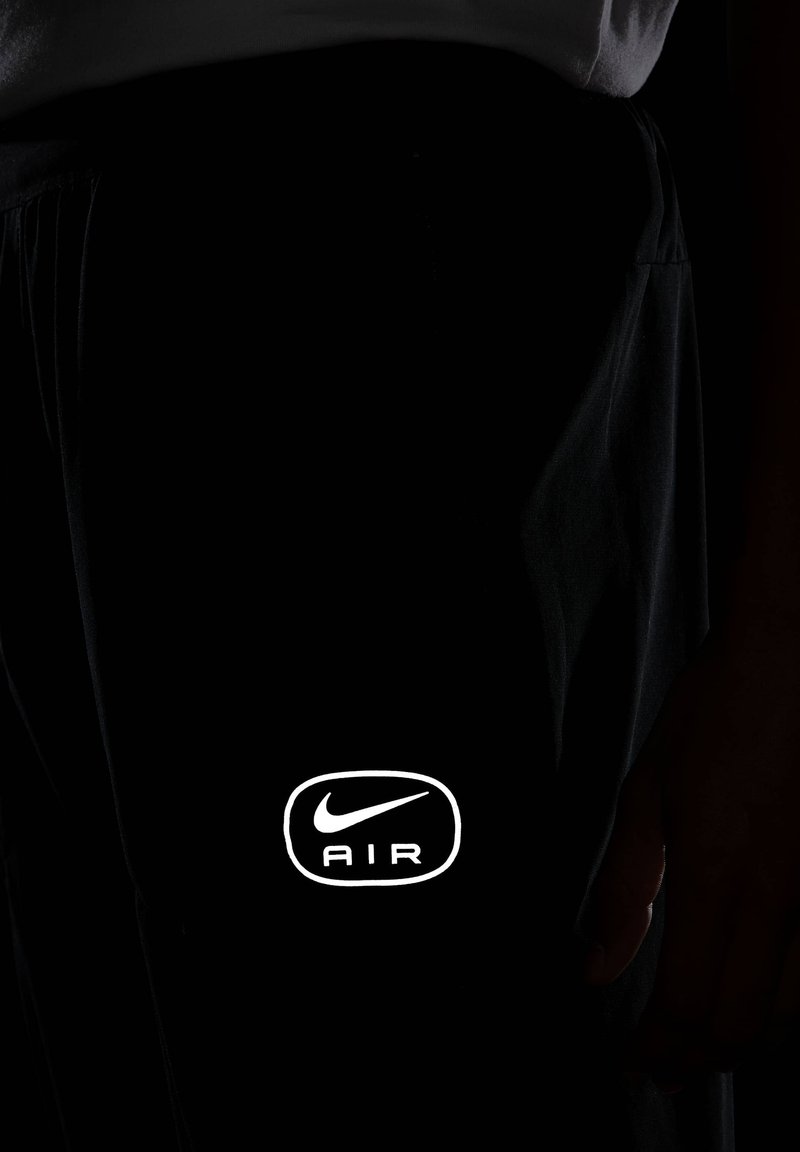 Un pantalon noir arbore un logo Nike blanc ainsi que le texte "AIR" sur le bas à gauche, avec une texture lisse et mate. Une main repose sur le tissu.