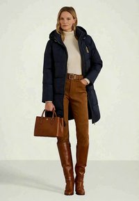 Moteris vilkintis tamsiai mėlyną gobtuvą turintį pūkinį paltą, kreminį megztinį su aukštu kaklu, rudus kelnius, iki kelių siekiančius rudus batus ir laikanti rudą odinę rankinę.