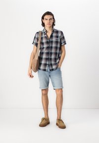 Cămașă scurtă cu mânecă în carouri în nuanțe de bleumarin, verde și alb, purtată cu șorturi din denim deschis rulat și pantofi din suede maro. Poartă o geantă de umăr bej.