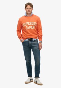 Oranje sweatshirt met de tekst "SUPERDRY JAPAN", blauwe overhemd eronder, blauwe spijkerbroek en witte sneakers met zwarte accenten.