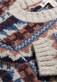 Artimiausiu planu vilnonis megztinis su daugiakampiu raštu mėlynos, rudos, kreminės ir tamsiai mėlynos spalvos atspalviais, atskleidžiantis apykaklės ir rankogalių detales.