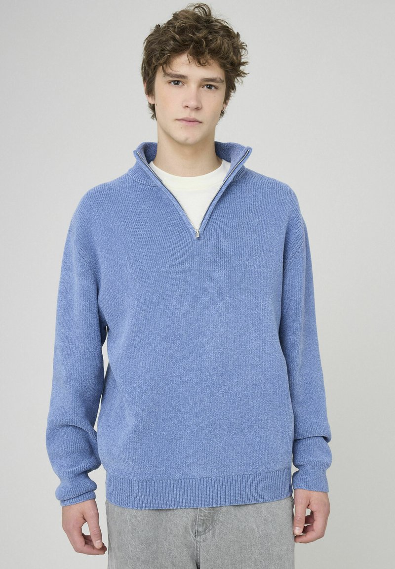 Pull en laine bleu clair avec un col zippé au quart, des poignets et un ourlet côtelés, et un motif de tricot texturé. Porté par-dessus une chemise blanche.