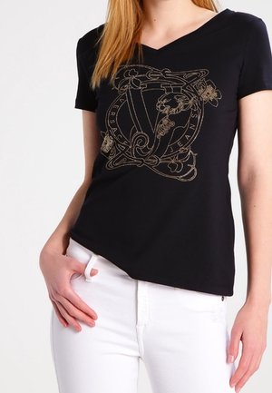 T-shirt noir à col en V avec un design graphique doré présentant des motifs complexes. Manches courtes, tissu doux, associé à un pantalon blanc.