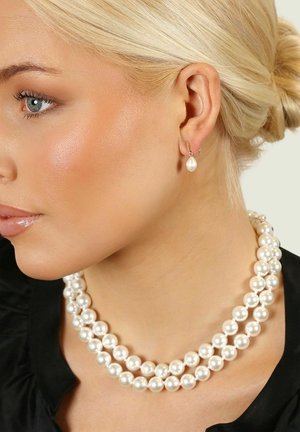 Tre strängar av stora vita pärlor bildar ett halsband. Ett matchande örhänge i form av en pärldropp bärs. Bakgrunden är neutral.