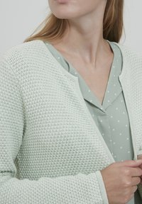 Cardigan lavorato a maglia verde chiaro con un motivo testurizzato, indossato sopra una camicetta verde menta a pois. Design con apertura frontale e bordi a costine.