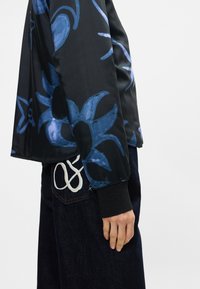 Veste noire avec motif floral bleu, tissu lisse, poignets côtelés et coupe décontractée, associée à un jean en denim foncé avec une couture décorative.