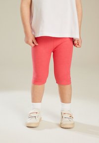 Koraalkleurige capri-legging van katoenmix, getailleerde stijl, naadloze zoom, gecombineerd met witte sokken en lichte schoenen met een regenboogaccent.