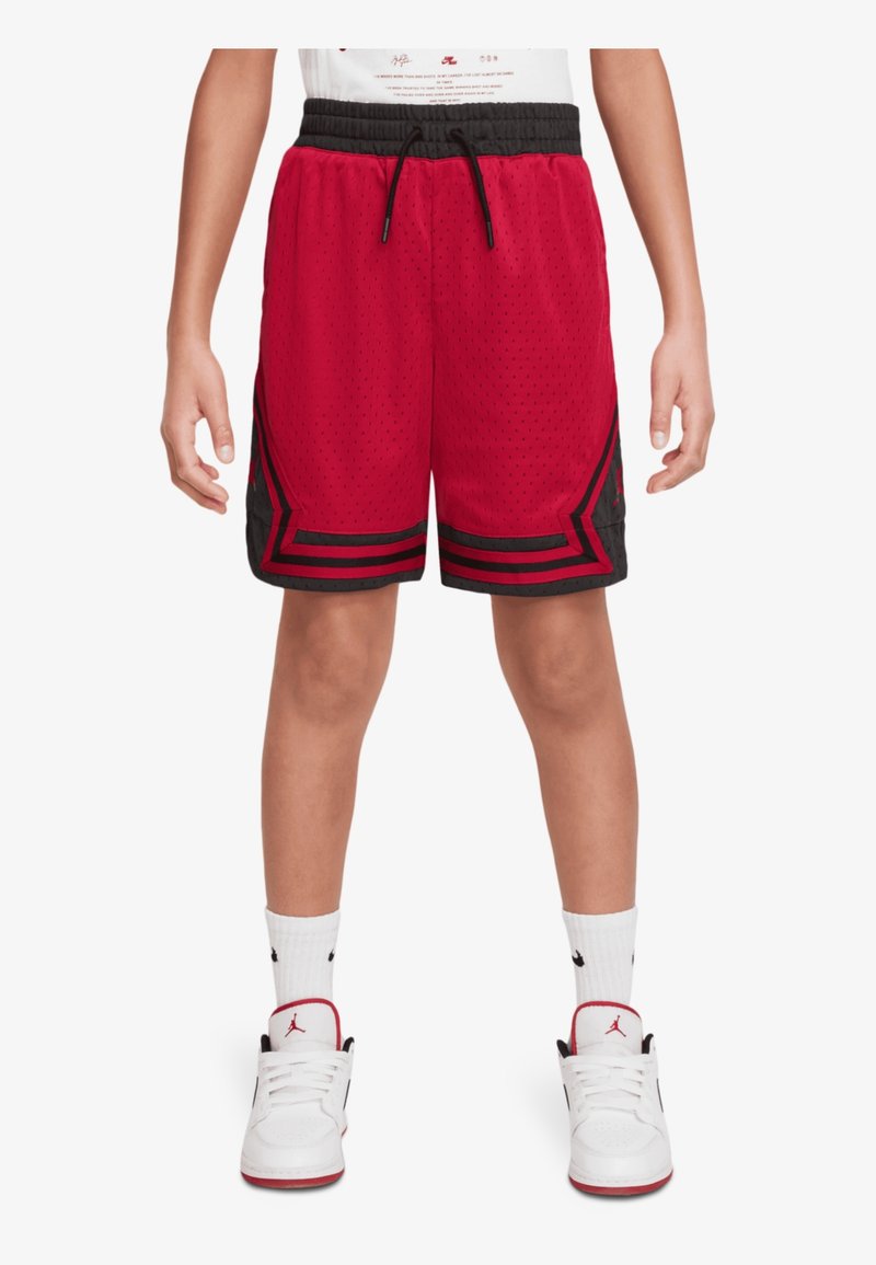 Enfant portant un short de basket rouge et noir, des chaussettes blanches et des baskets blanches, debout les jambes légèrement écartées.