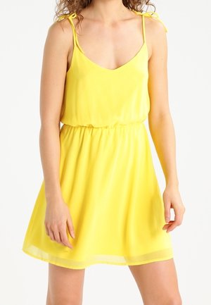 Femme portant une robe jaune sans manches avec des bretelles nouées et une taille élastique, debout devant un fond blanc.