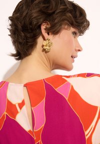 Kolorowa sukienka w wzory z żywymi odcieniami różu, pomarańczy i kremu; z dekoltem w kształcie dziurki oraz złotą biżuterią w kształcie kwiatu.