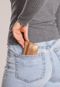 Cilindro di metallo dorato con zip parzialmente inserito in una tasca posteriore di jeans azzurri, con una superficie testurizzata e forma arrotondata. Top grigio a maniche lunghe visibile.