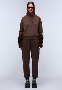 Felpa con cappuccio marrone, dotata di tasca frontale e polsini larghi a coste, abbinata a pantaloni jogger coordinati con tasche laterali e polsini elasticizzati.