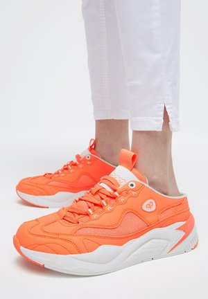 Helle orange Sneakers mit weißen Sohlen, getragen zu weißen, gekürzten Hosen, die Gelenk- und Schuhdetails vor schlichtem Hintergrund zeigen.