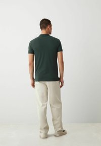 Camisa polo de color verde oscuro con mangas cortas, combinada con pantalones de color crema y zapatillas beige, mostrando un estilo sencillo y informal.