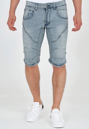 Mann iført lyseblå denim shorts til kneet med dekorative sømmer og hvite joggesko, stående med avslappede hender langs siden.