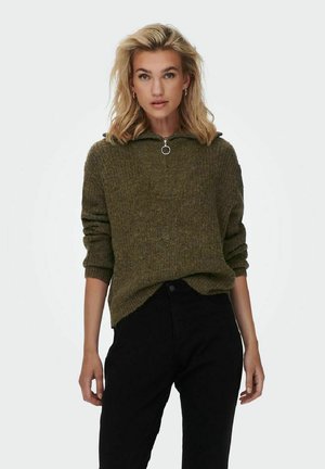 Maglione lavorato a maglia verde oliva con collo alto, dettaglio con zip e maniche arrotolate. Abbinato a pantaloni in denim neri, evidenziando un look casual.