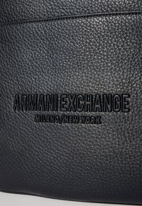 Prodotto in pelle nera testurizzata con logo in rilievo "ARMANI EXCHANGE MILANO/NEW YORK". La superficie presenta una finitura a ciottoli.