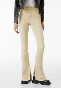 Beiga utsvängda jeans med sidofickor, tillverkade av denim. Har framfickor, knäppning med knapp och en åtsittande midja. Svarta platåstövlar bärs.