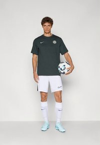 Tamnozelena Nike majica s logotipom, bijele Nike hlače, bijele čarape s plavim detaljima i svijetloplave kopačke. Drži nogometnu loptu s uzorkom.