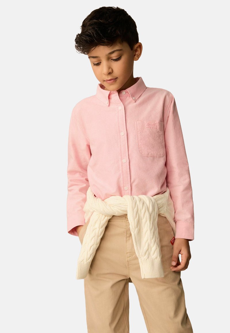 Camicia button-up rosa chiaro con taschino, abbinata a pantaloni beige. Un maglione in maglia cable-knit color crema è annodato in vita.