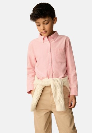Garçon portant une chemise rose clair à boutons, un pull crème torsadé noué autour de la taille et un pantalon beige, regardant vers le bas.