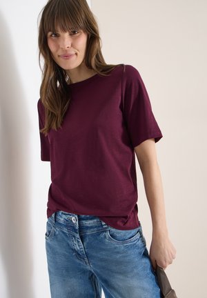 Femme aux cheveux bruns portant un t-shirt bordeaux à manches courtes et un jean bleu, tenant un sac à main marron sur un fond neutre.