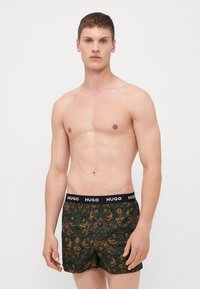 Boxershorts aus dunkelgrünem Stoff mit floralem und vogelartigem Muster. Der Bund ist schwarz mit weißem "HUGO"-Logo.
