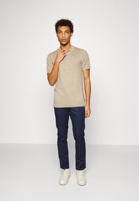 Beige polo shirt med korta ärmar, matchat med marinblå byxor och vita sneakers. Enkel design med en diskret logodetalj på bröstet.