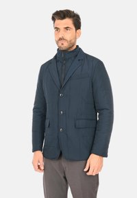 Blazer trapuntato blu navy con colletto alto, chiusura a bottoni singola e due tasche a toppa davanti. Texture liscia.