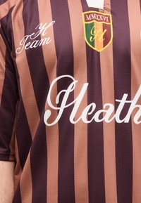 Αθλητική φανέλα με κάθετες ρίγες σε καφέ και σκούρο μοβ χρώμα, με λευκό κείμενο "Heath", κίτρινη και πράσινη σημαία, και τη φράση "Y Team" στο στήθος.