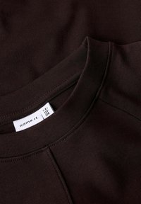 Tuum brown crewneck vaatte, jossa on näkyvät saumat ja valkoinen vaatetunnus, jossa näkyy brändin nimi ja koko.