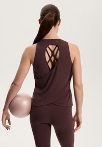 Bordeauxfarbenes Sporttanktop mit einem Kreuzrücken-Design, kombiniert mit passenden Leggings, gehalten von einer Person neben einem hellrosa Übungsball.