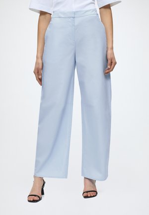 Personne portant un pantalon large bleu clair, un haut blanc et des sandales à talons noires ouvertes à l'avant, debout avec les bras détendus le long du corps.