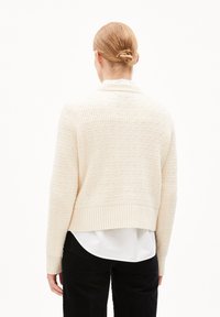 Cr�ème-kleurige gebreide cardigan met een gestructureerd patroon, geribbelde zoom en manchetten, gezien vanaf de achterkant over een wit shirt en zwarte broek.