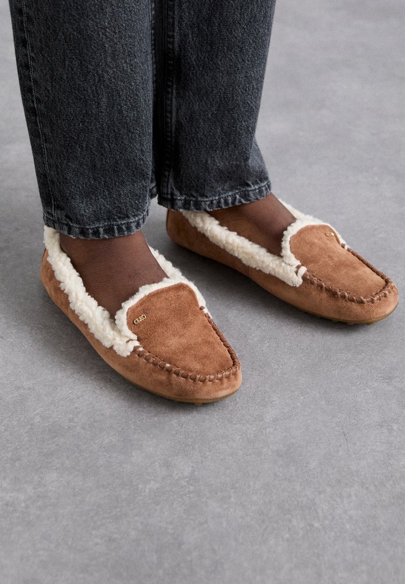 Pantoufles en suède brun avec doublure en shearling blanc, au design classique de mocassin, présentant des détails cousus à la main et un petit accent en métal logo.