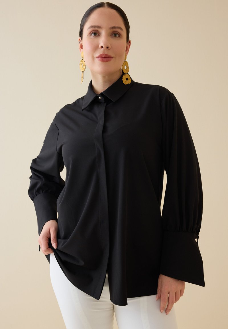 Mujer con blusa negra de manga larga con grandes puños y pendientes dorados colgantes, posando frente a un fondo beige.