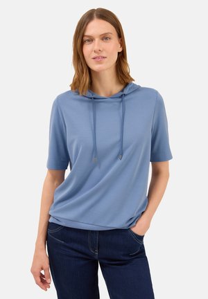Une jeune femme aux cheveux bruns mi-longs porte un hoodie à manches courtes bleu clair et un jean bleu foncé, se tenant avec une main dans la poche.