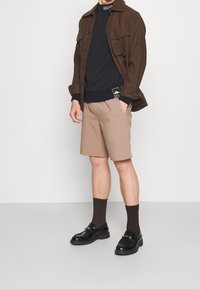 Chaqueta marrón de cuadros, camisa negra, pantalones cortos beige, calcetas marrones oscuros, mocasines negros con herrajes dorados. Enfócate en el uso casual de capas y los detalles de textura.