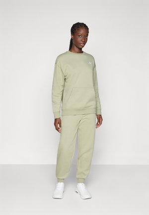 Lys grøn sweater og matchende bukser lavet af blødt stof, med en frontlomme og et lille logo på brystet. Hvide sneakers.