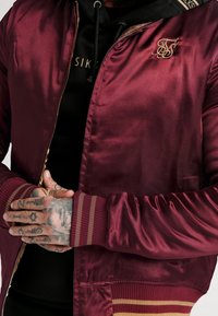 Burgundy satin bomberjakke med ribbede manchetter med røde og gyldne striber, lynlåslukning og et broderet logo på brystet.
